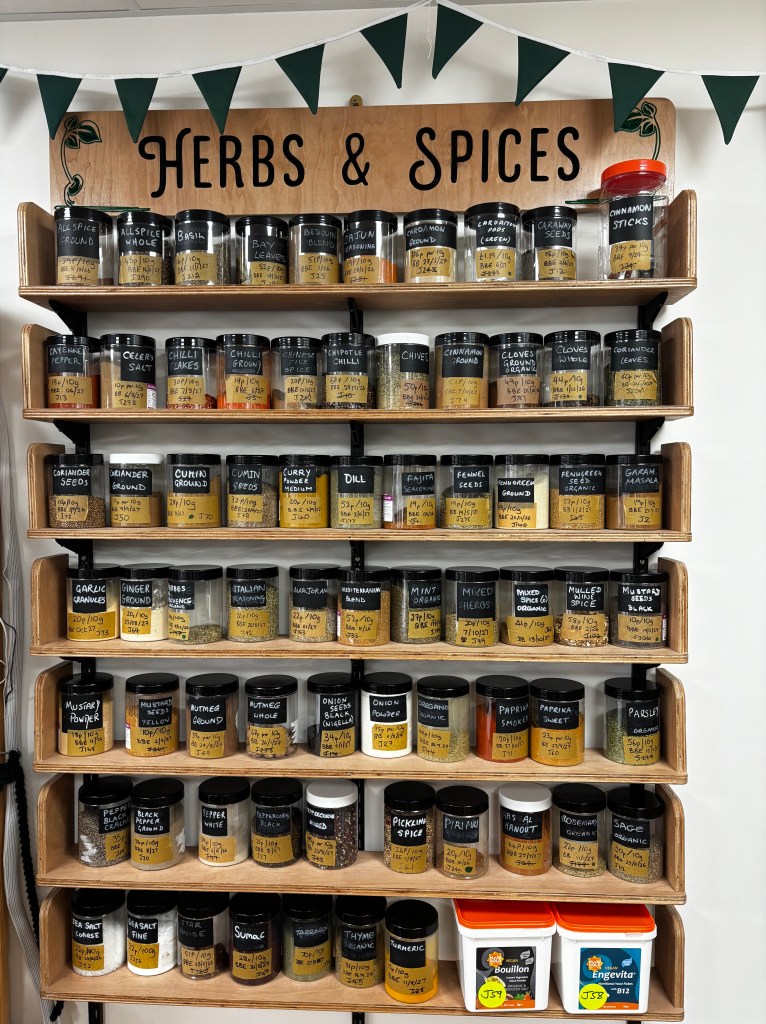 Herbs and spices display at The Green Gram with rows of refill jars labelled by hand, offering a wide range of dried herbs, spices, blends, and seasonings.