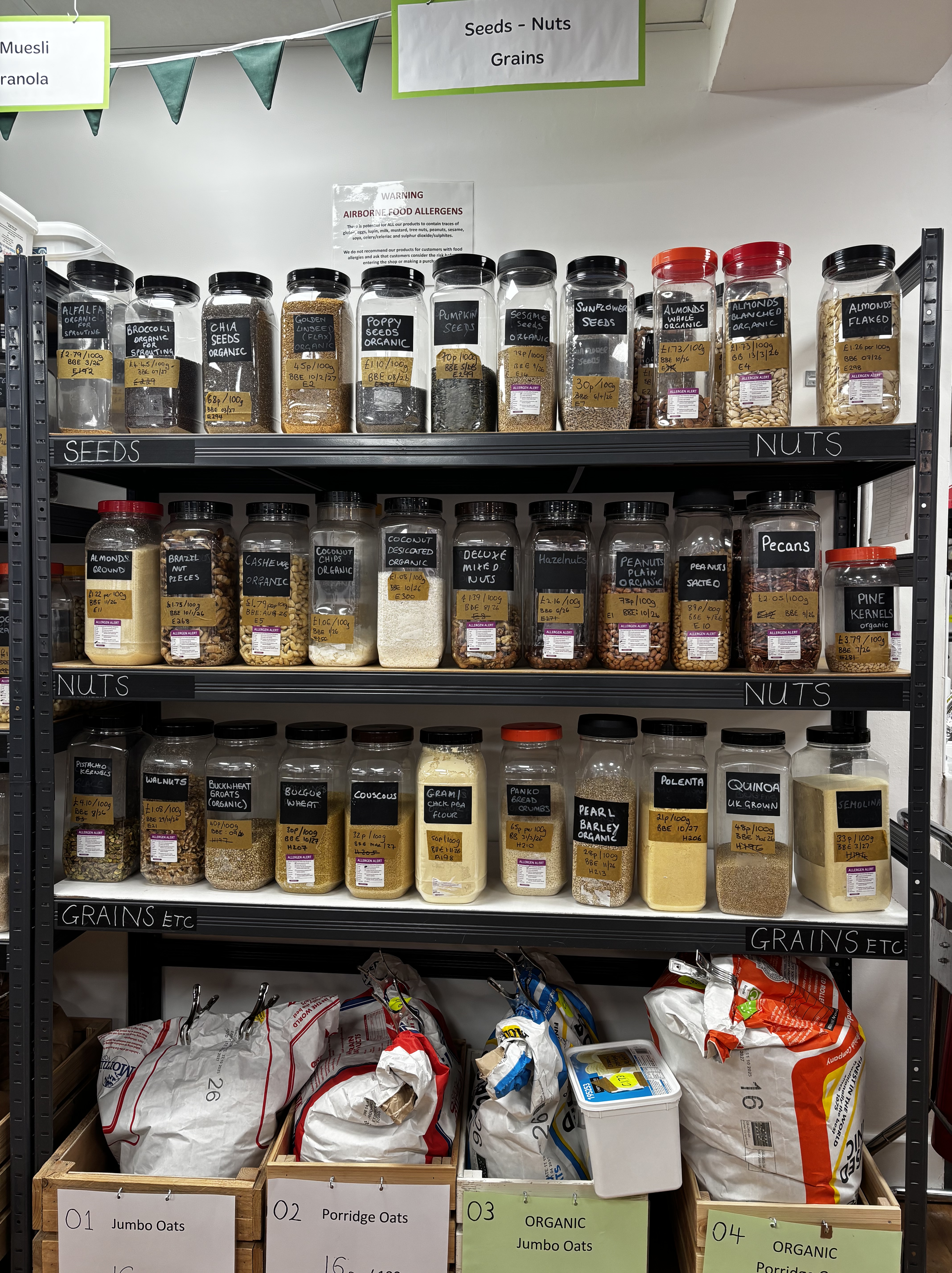 Seeds, nuts, and grains refill shelves at The Green Gram, showing large labelled jars of nuts, seeds, oats, and pantry staples, with bulk sacks stored below.