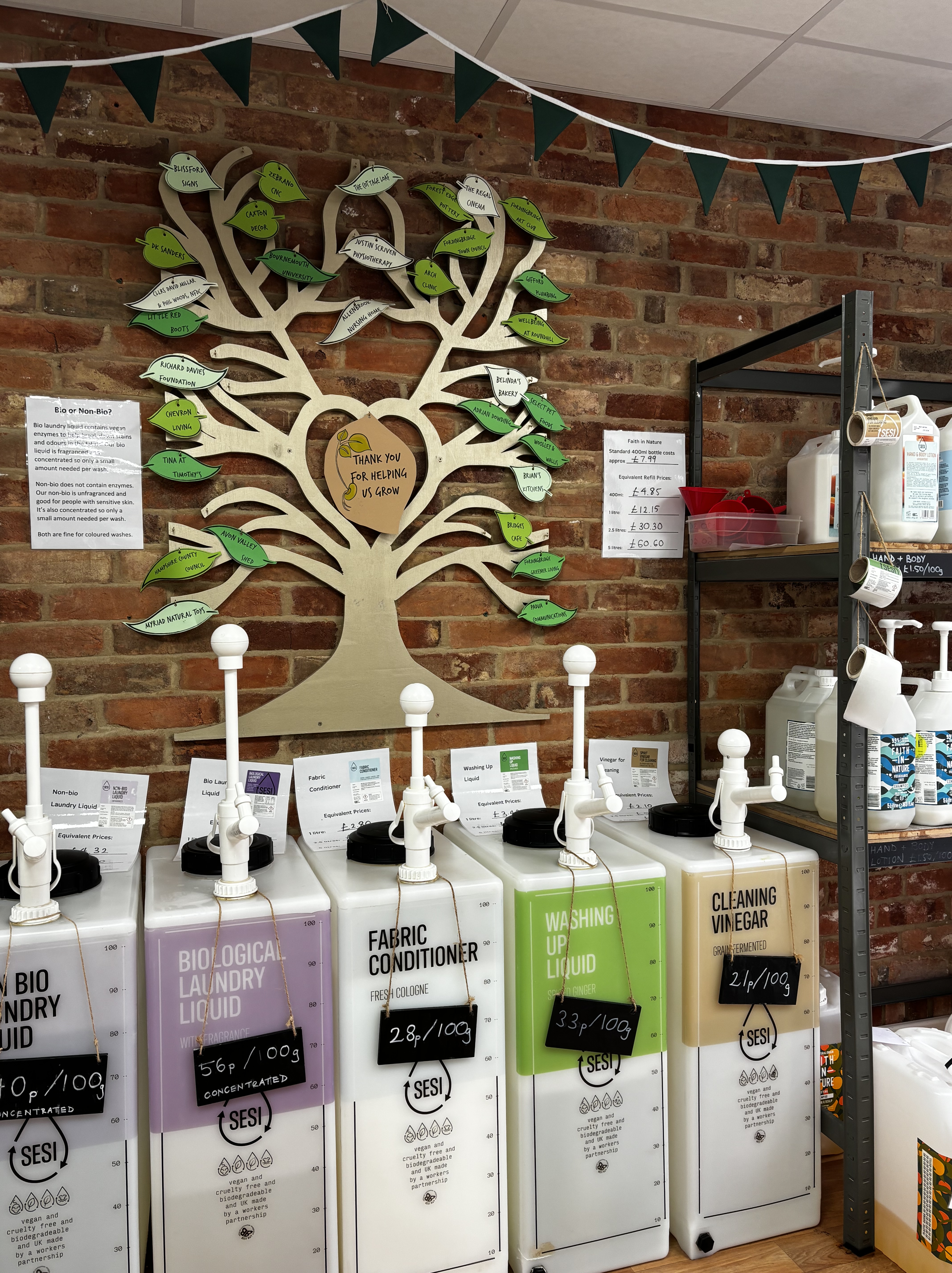 Refill station at The Green Gram with SESI household liquid dispensers for laundry, conditioner, washing up liquid, and cleaning vinegar, set against a brick wall with a wooden thank you tree decorated with supporter names.