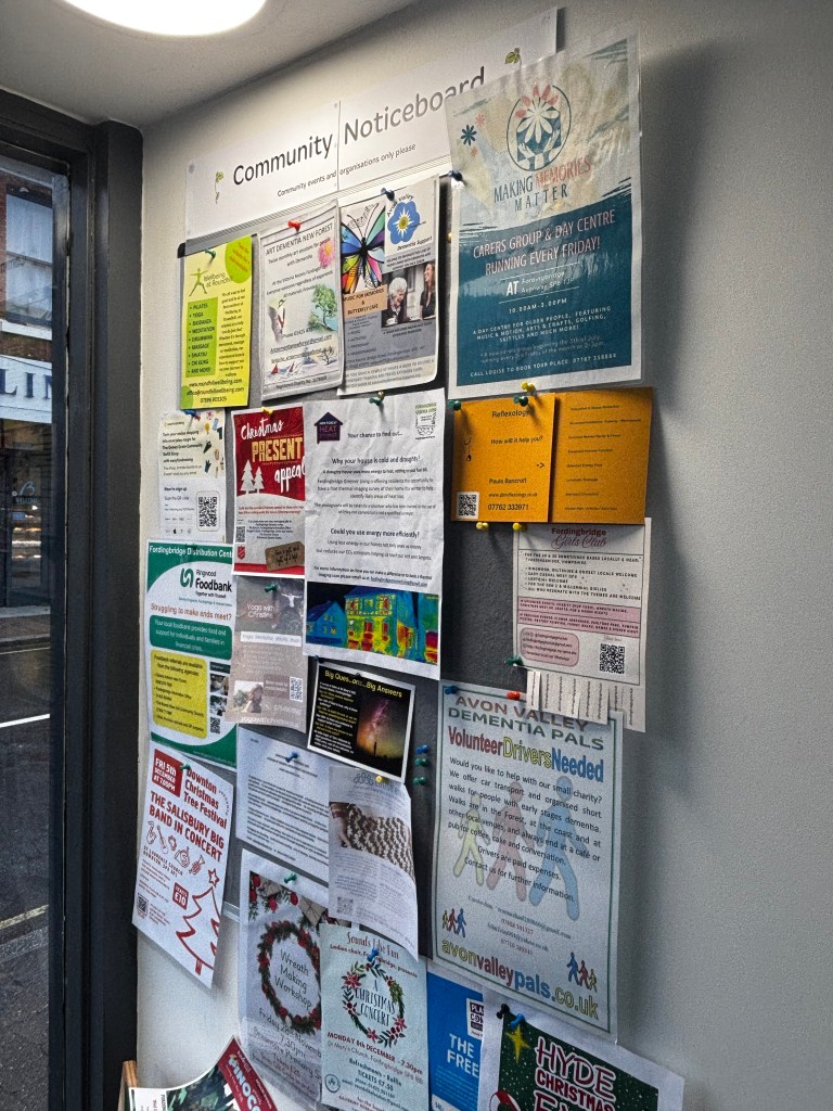 Community noticeboard near the entrance of The Green Gram filled with local event posters, charity appeals, workshops, and information for residents.
